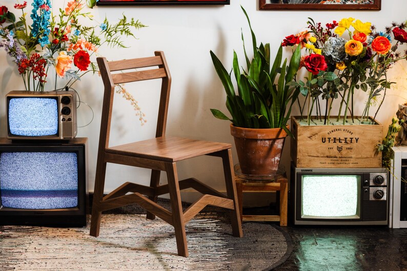 May include: A wooden chair with a unique angled leg design stands in front of a wall. Two vintage televisions with static screens are on the floor. Colorful flowers are arranged in vases and wooden crates, adding a vibrant touch to the scene.