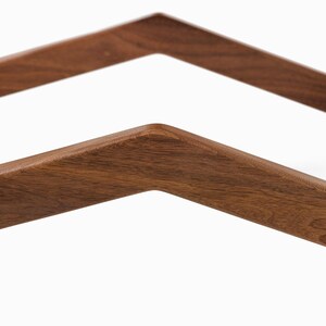 May include: Close-up of a wooden furniture frame with a geometric design. The frame is made of dark brown wood and features angled, V-shaped supports. The wood grain is visible, and the edges are rounded.
