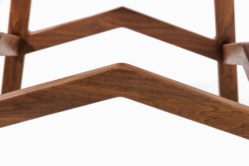 May include: Close-up of a wooden furniture frame with a geometric design. The frame is made of dark brown wood and features angled, V-shaped supports. The wood grain is visible, and the edges are rounded.