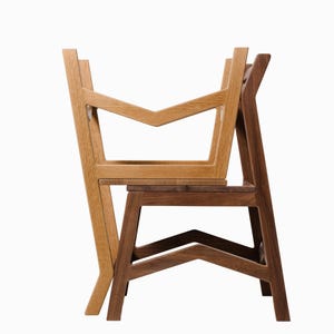 May include: Two wooden chairs, one light-colored and one dark-colored, are stacked against each other. The chairs have a modern design with geometric shapes and angled legs. The light-colored chair is in front, and the dark-colored chair is behind.