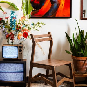 May include: A wooden chair with a minimalist design stands in a room. The chair has a rectangular seat and a back with two horizontal slats. A vintage television set is in the background, along with colorful flowers and a potted plant.