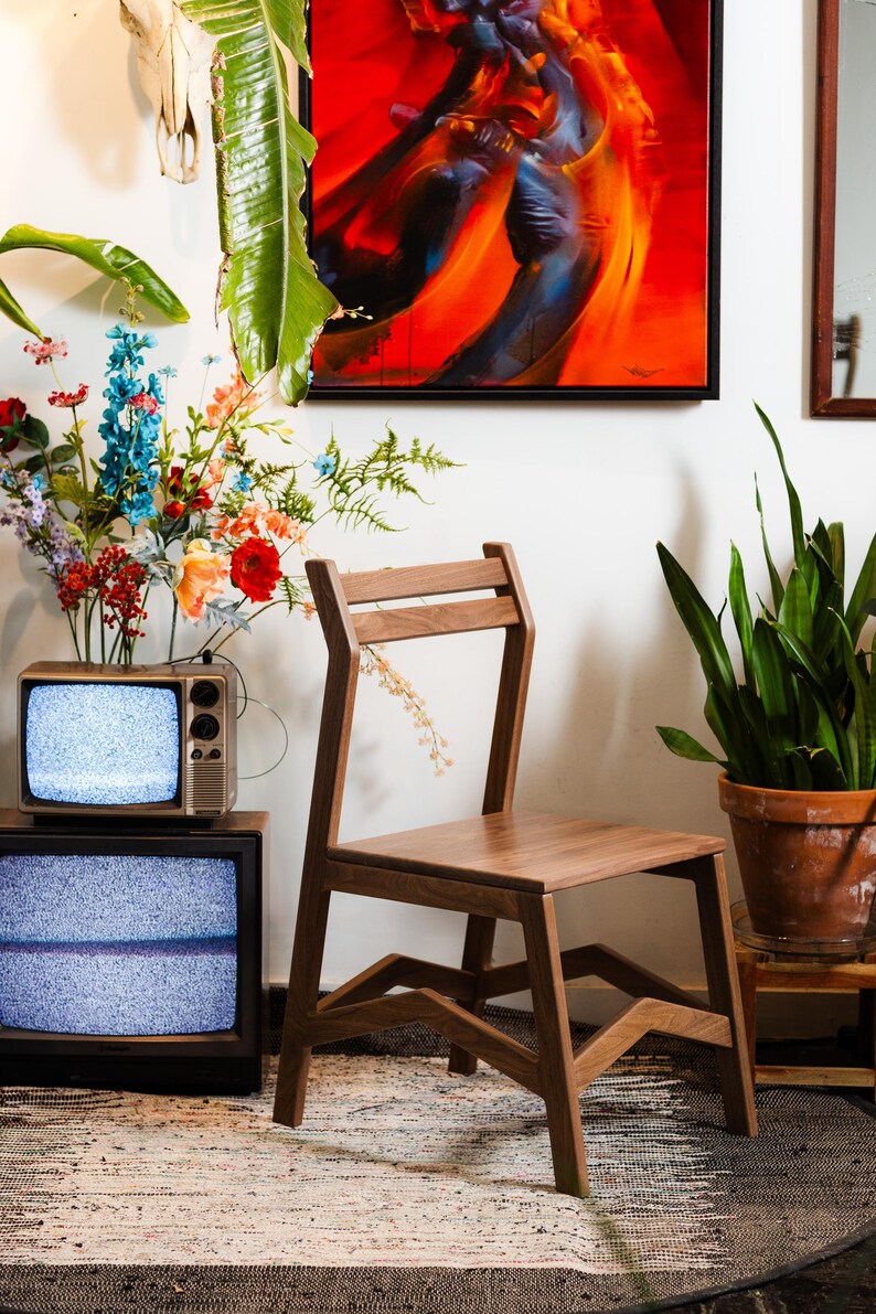 May include: A wooden chair with a minimalist design stands in a room. The chair has a rectangular seat and a back with two horizontal slats. A vintage television set is in the background, along with colorful flowers and a potted plant.
