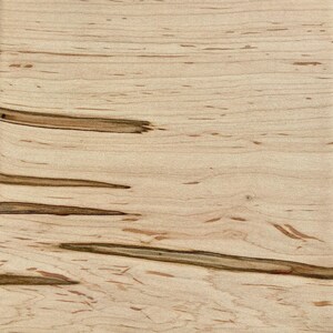 May include: Close-up of a light-colored wood grain pattern. The wood has a natural, light tan base color with darker brown streaks and small, scattered dark spots. The grain runs horizontally across the image.
