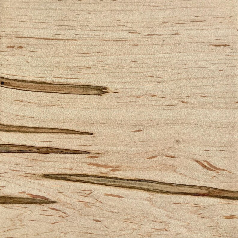 May include: Close-up of a light-colored wood grain pattern. The wood has a natural, light tan base color with darker brown streaks and small, scattered dark spots. The grain runs horizontally across the image.