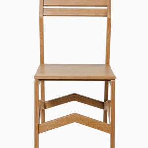 May include: A light-colored wooden chair with a rectangular seat and a backrest featuring two horizontal slats. The chair has four legs and a distinctive V-shaped support structure. The chair is made of wood.