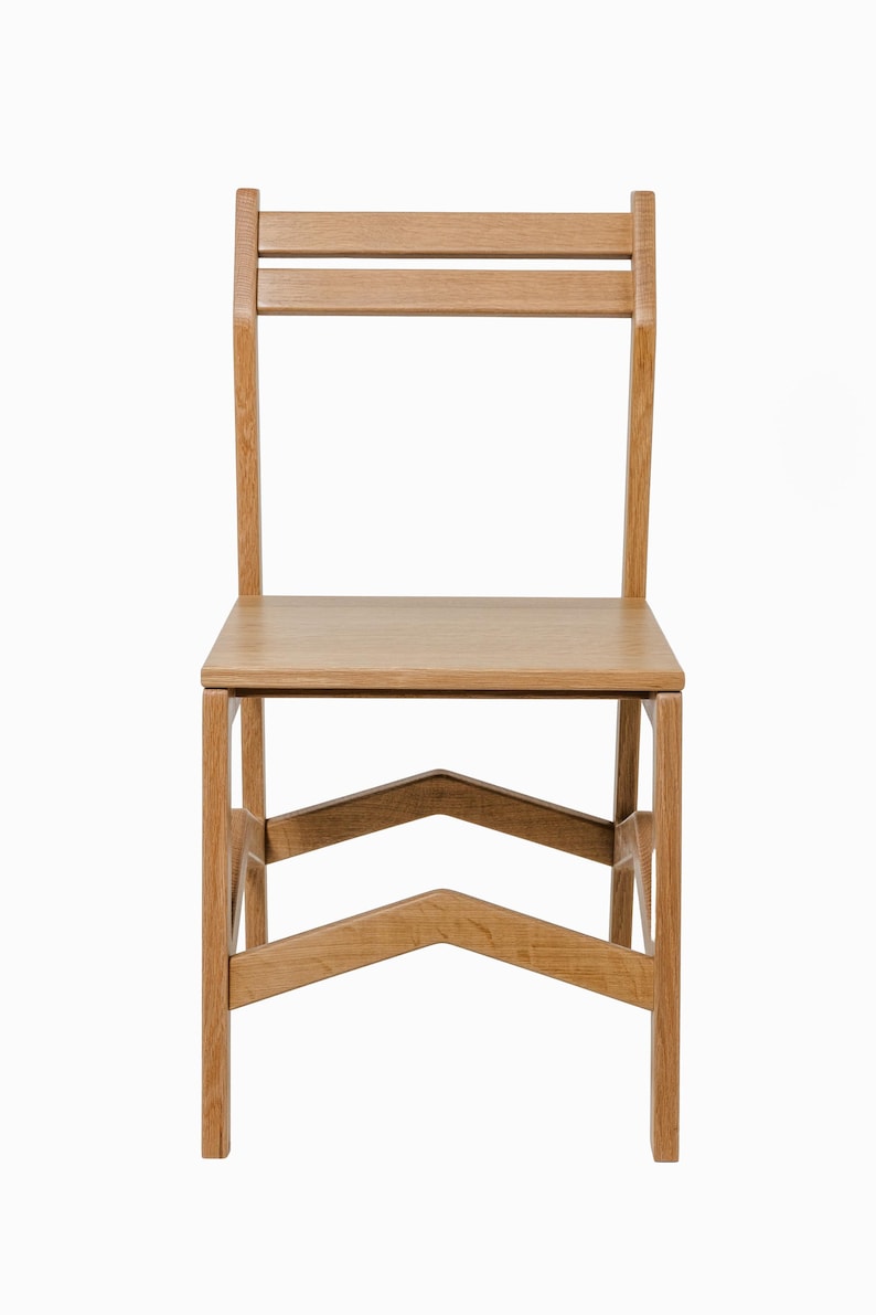May include: A light-colored wooden chair with a rectangular seat and a backrest featuring two horizontal slats. The chair has four legs and a distinctive V-shaped support structure. The chair is made of wood.