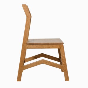 May include: A side view of a wooden chair with a light brown finish. The chair has a straight back with a curved top, a rectangular seat, and a geometric base. The chair's dimensions are approximately 36 inches high, 18 inches wide, and 18 inches deep.