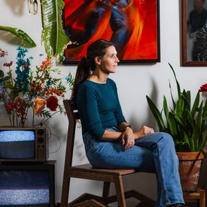 May include: A woman sits on a wooden chair, wearing a teal top and blue jeans. Behind her is a vibrant abstract painting with red and orange hues. A vintage television set and potted plants are also visible.