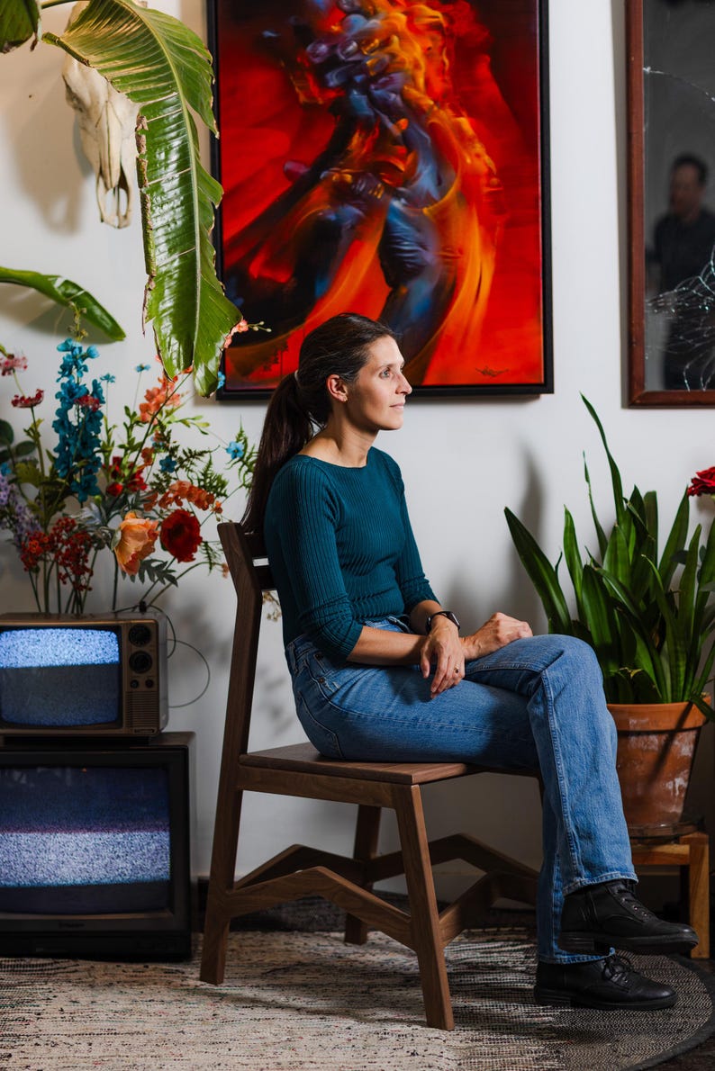 May include: A woman sits on a wooden chair, wearing a teal top and blue jeans. Behind her is a vibrant abstract painting with red and orange hues. A vintage television set and potted plants are also visible.