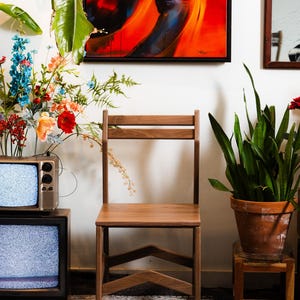 May include: A wooden chair with a simple design stands in front of a white wall. A colorful abstract painting hangs above the chair. A vintage television and potted plants are also in the room.