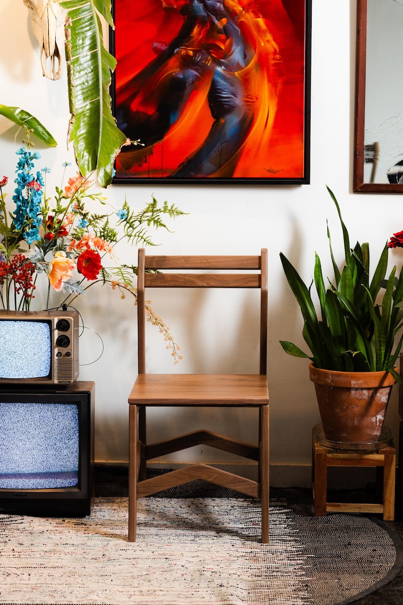 May include: A wooden chair with a simple design stands in front of a white wall. A colorful abstract painting hangs above the chair. A vintage television and potted plants are also in the room.