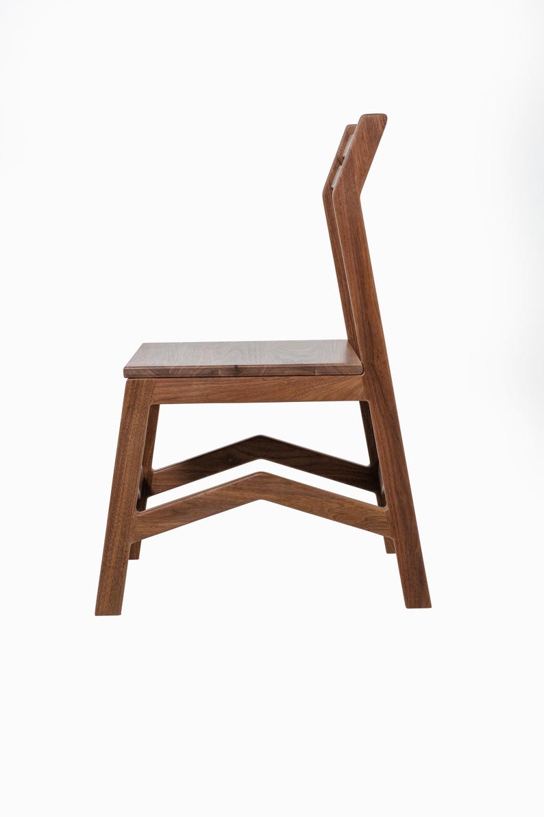 May include: A side view of a wooden chair with a minimalist design. The chair is constructed from dark brown wood, featuring a rectangular seat and a tall backrest with a unique, angled design. The legs are sturdy and angled, with a V-shaped support.