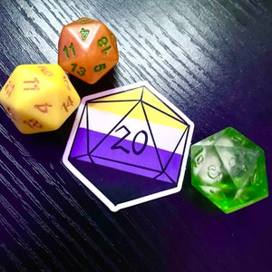Può includere: Un adesivo di un dado a 20 facce con i colori della bandiera del pride non binario. Il dado è giallo, bianco e viola. Nell'immagine ci sono tre dadi, uno giallo, uno arancione e uno verde.