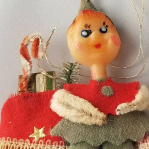 May include: A vintage Christmas ornament depicting a doll-like figure with a plastic face, red felt outfit, and green felt trousers. The ornament has a gold star and lace trim. A candy cane and small gift box are attached.