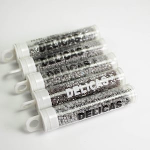May include: Five clear plastic cylindrical tubes filled with small grey beads. Each tube features a white cap with a hanging loop and black text printed on the side that reads DELICAS, MIYUKI SHOJI, and OPAQUE MATTE RAINBOW PALE GREY.
