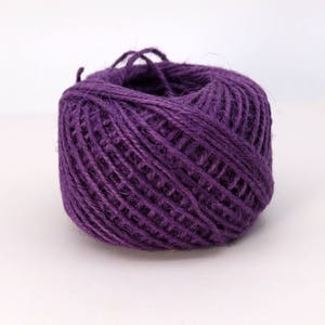May include: A round ball of purple jute twine featuring a thick, fibrous texture with visible strands wound tightly together. The twine has a consistent deep violet hue throughout, with loose ends extending from the top of the spherical spool.