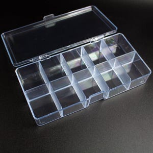 May include: A clear plastic storage box featuring a hinged lid and ten individual rectangular compartments arranged in two rows of five. The container is constructed from transparent material, allowing for full visibility of the interior sections.
