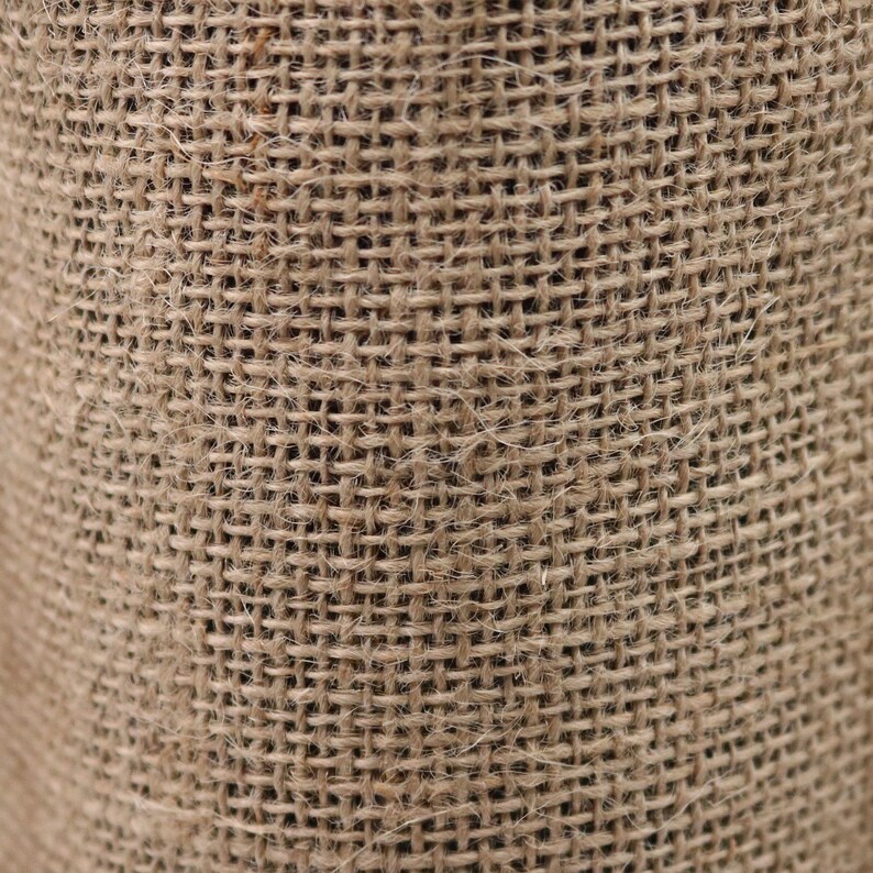 May include: A close-up view of a coarse, tan-colored burlap fabric featuring a distinct, open-weave grid pattern. The material displays a natural, fibrous texture with visible individual strands of jute woven together in a simple, uniform crisscross design.