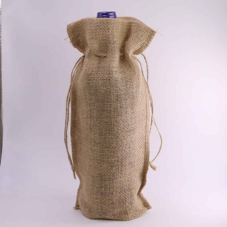 May include: A tan burlap wine bottle gift bag featuring a coarse woven texture, a drawstring closure at the top with matching twine cords, and a visible purple bottle cap protruding from the opening of the fabric container.