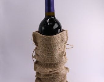 May include: A dark glass wine bottle with a purple foil seal on the neck, partially encased in a tan, woven hessian drawstring bag. The coarse, textured fabric of the bag bunches around the lower portion of the bottle, featuring visible twine.