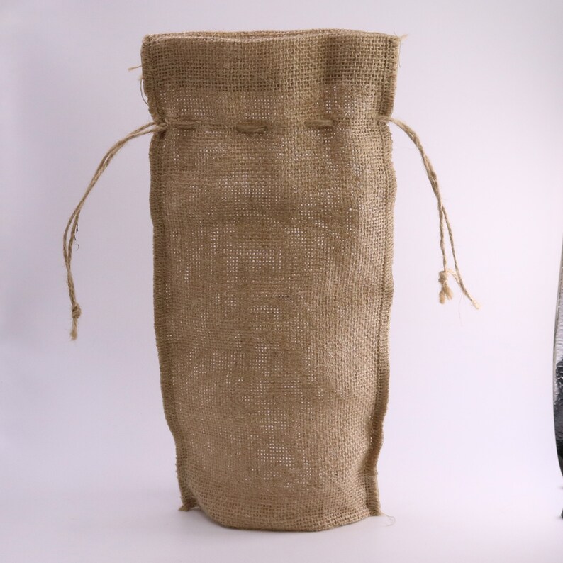 May include: A tall, rectangular drawstring bag crafted from natural tan-colored burlap fabric. The bag features a coarse, woven texture and includes two thin, matching jute strings threaded through the top hem for closure and carrying purposes.