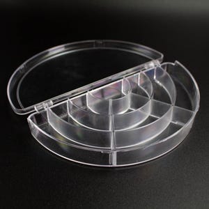 Puede incluir: Un recipiente de almacenamiento de plástico transparente en forma de semicírculo con tapa abatible y múltiples compartimentos internos dispuestos en arcos concéntricos. El organizador tiene un acabado claro y un diseño estructurado.