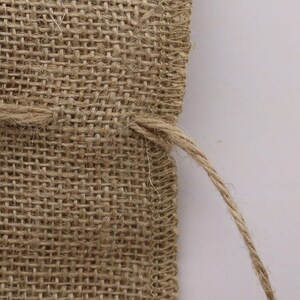May include: A close-up view of a tan burlap fabric pouch featuring a coarse, woven texture with visible fibers. The edges are finished with a simple stitched seam, and a thin, matching jute drawstring cord extends from the side of the pouch.