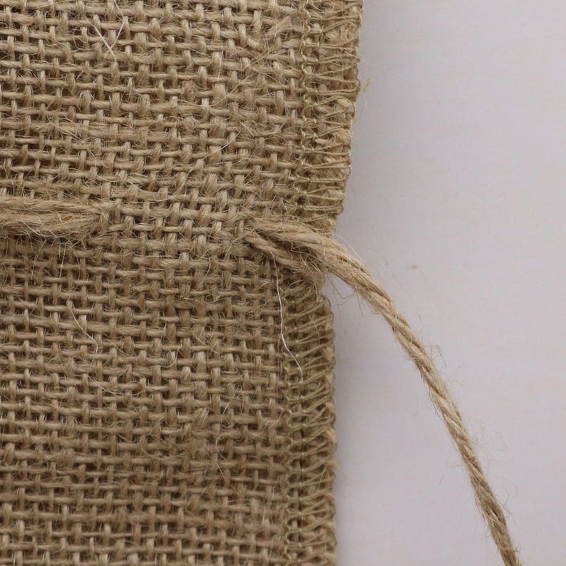 May include: A close-up view of a tan burlap fabric pouch featuring a coarse, woven texture with visible fibers. The edges are finished with a simple stitched seam, and a thin, matching jute drawstring cord extends from the side of the pouch.