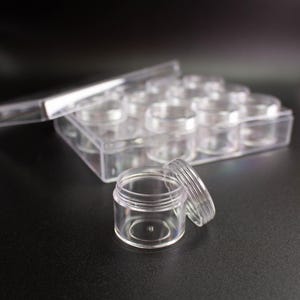 Pode incluir: Um recipiente de armazenamento de plástico transparente com vários frascos pequenos e redondos de tampa roscada dispostos em grade. Um frasco vazio individual com a tampa a condizer está em primeiro plano, separado da caixa principal.