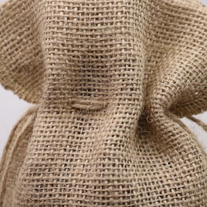 May include: A tan burlap drawstring bag featuring a coarse, woven texture with visible fibers. The fabric has a distinct grid-like pattern created by the interlacing of thick, natural-colored threads, forming a durable and rustic material structure.