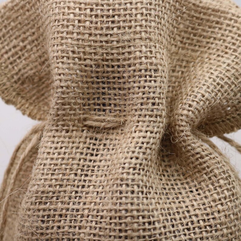 May include: A tan burlap drawstring bag featuring a coarse, woven texture with visible fibers. The fabric has a distinct grid-like pattern created by the interlacing of thick, natural-colored threads, forming a durable and rustic material structure.