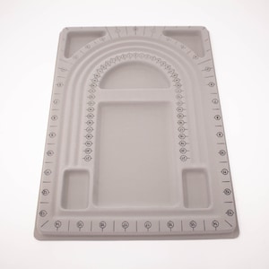 May include: A light gray rectangular jewelry design board featuring a flocked surface with U-shaped channels for necklace layout. The board includes printed measurement markings, numbered circles from 1 to 21 along the channels, and recessed compartments for organizing beads and jewelry components.