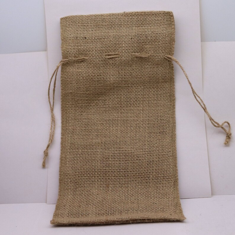 May include: A rectangular drawstring pouch crafted from tan-colored, coarse-woven burlap fabric. The bag features a simple top closure mechanism with two thin, matching jute strings extending from the sides, allowing for easy opening and closing.