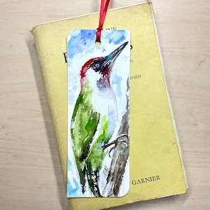 Peut inclure: Un marque-page rectangulaire en papier présentant une peinture à l'aquarelle d'un pic vert avec une calotte rouge, perché sur une branche d'arbre brune sur fond de ciel bleu, agrémenté d'un fin ruban rouge noué dans un trou au sommet.