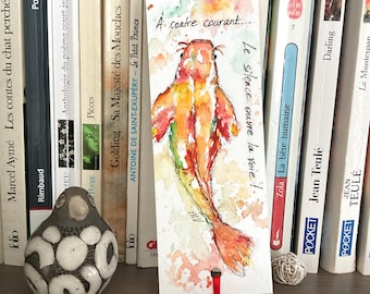 May include: A rectangular paper bookmark featuring a watercolor painting of a koi fish in shades of orange, red, and yellow. The text "À contre courant... Le silence ouvre la voie!" is written in black ink. A red ribbon is attached to the bottom hole.