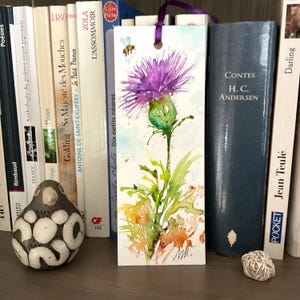 May include: Watercolor bookmark featuring a purple thistle flower with green leaves and a small bee. The bookmark has a purple ribbon and the text "Dix petits nêares". The bookmark is displayed among books.