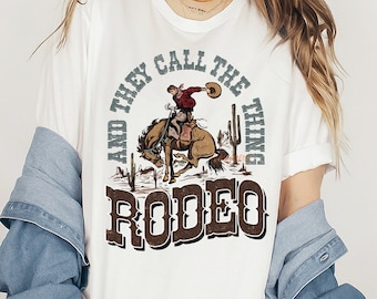 May include: White t-shirt with a graphic design of a cowboy riding a bucking horse. The text reads "And they call the thing rodeo".