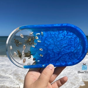 Op de afbeelding: Een blauwe harsbak met een helder gedeelte gevuld met schelpen en kleine metalen zeedieren. De bak is ovaalvormig en heeft een getextureerd oppervlak.