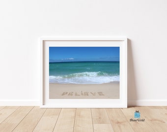 May include: A framed photograph of a beach scene with the word "BELIEVE" written in the sand. The ocean is a bright turquoise blue with white waves crashing on the shore. The sky is a clear blue with a few white clouds.