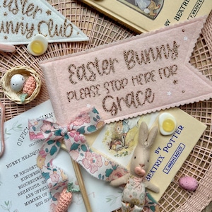 May include: An Easter-themed display featuring a pink banner with glitter text saying "Easter Bunny PLEASE STOP HERE FOR Grace", a bunny doll, and a Beatrix Potter book. Also includes decorative eggs.