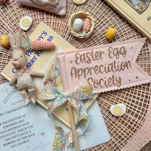 May include: An Easter-themed arrangement with a plush bunny, miniature carrots, and a banner reading "Easter Egg Appreciation Society." The scene includes a small basket with eggs, a book, and decorative eggs, all arranged on a pink woven mat.
