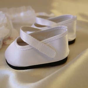 May include: A pair of white satin baby shoes with a strap and buckle fastening. The shoes have a black sole.