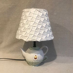 Könnte beinhalten: Dekorative Tischlampe mit weißem, plissiertem Lampenschirm auf einem teekannenförmigen Sockel. Der Sockel ist weiß mit hellgrüner Zierleiste und einer Cupcake-Illustration. Die Lampe hat ein schwarzes Netzkabel.