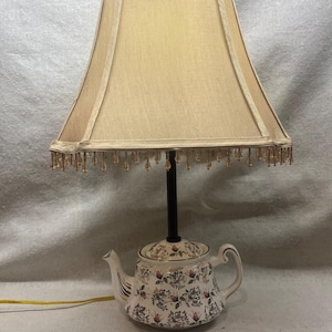 May include: A table lamp featuring a white ceramic teapot base decorated with a floral pattern of pink rosebuds and green leaves. The lamp has a black central stem and a beige fabric lampshade with a decorative beaded fringe along the bottom edge.