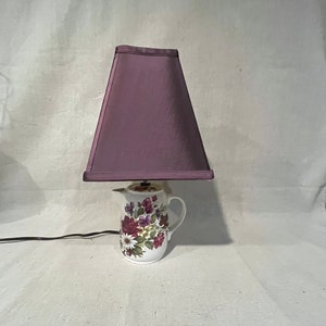 Può includere: Lampada decorativa con paralume viola su una brocca in ceramica a motivi floreali. La brocca è bianca con un disegno di fiori rossi, viola e gialli. Un cavo di alimentazione nero si estende dalla base.