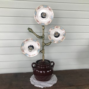 May include: A unique lamp made from repurposed materials. It features three vintage plates with floral designs, each holding a light bulb. The base is a brown ceramic crock pot.
