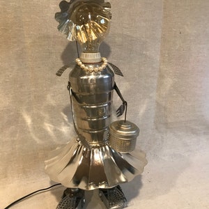 May include: A quirky, handmade lamp made from recycled metal parts. The lamp has a lightbulb head, a pearl necklace, and a skirt-like design. It holds a small basket and has metal feet. The lamp is plugged in with a black cord.