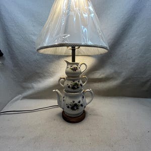 Könnte beinhalten: Eine Tischlampe mit einem Fuß aus einer gestapelten weißen Porzellanteekanne, Zuckerdose und Milchkännchen, die mit violetten Blumenmustern verziert sind. Die Lampe steht auf einem runden Holzsockel und hat einen mit Kunststoff bedeckten weißen Lampenschirm.