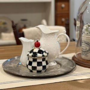 May include: A decorative arrangement featuring a silver tray holding a small white pitcher, a white dotted pitcher, and a black and white checkered jar with a red cherry-like top. The items are on a wooden table.