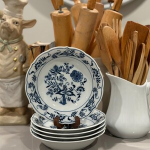 May include: A stack of white porcelain bowls with a blue floral design. The bowls have a scalloped edge and are stacked on a wooden stand. A white pitcher and wooden kitchen utensils are in the background.